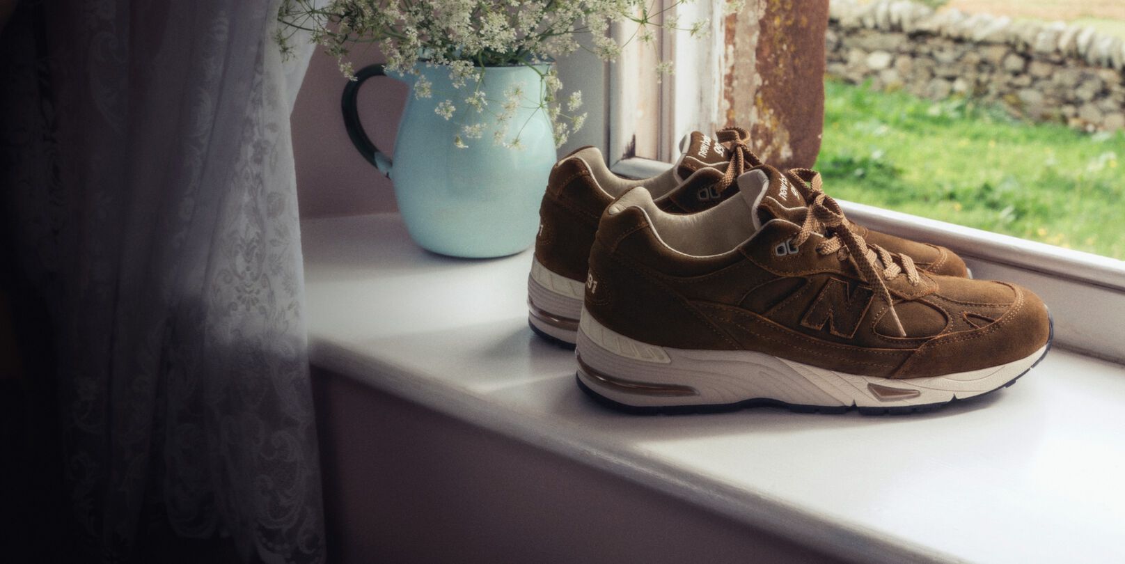 A pair of New Balance Made in UK sneakers on a windowsill next to a small vase of white flowers. 
