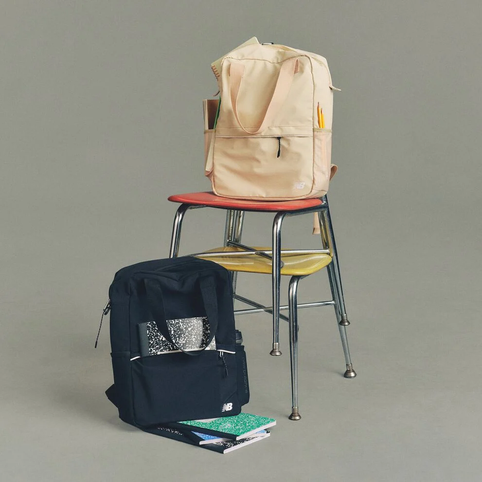 Studio image of a pair of cream-colored New Balance sneakers placed on a dark wooden folding chair, with a second matching chair folded and leaned behind it.