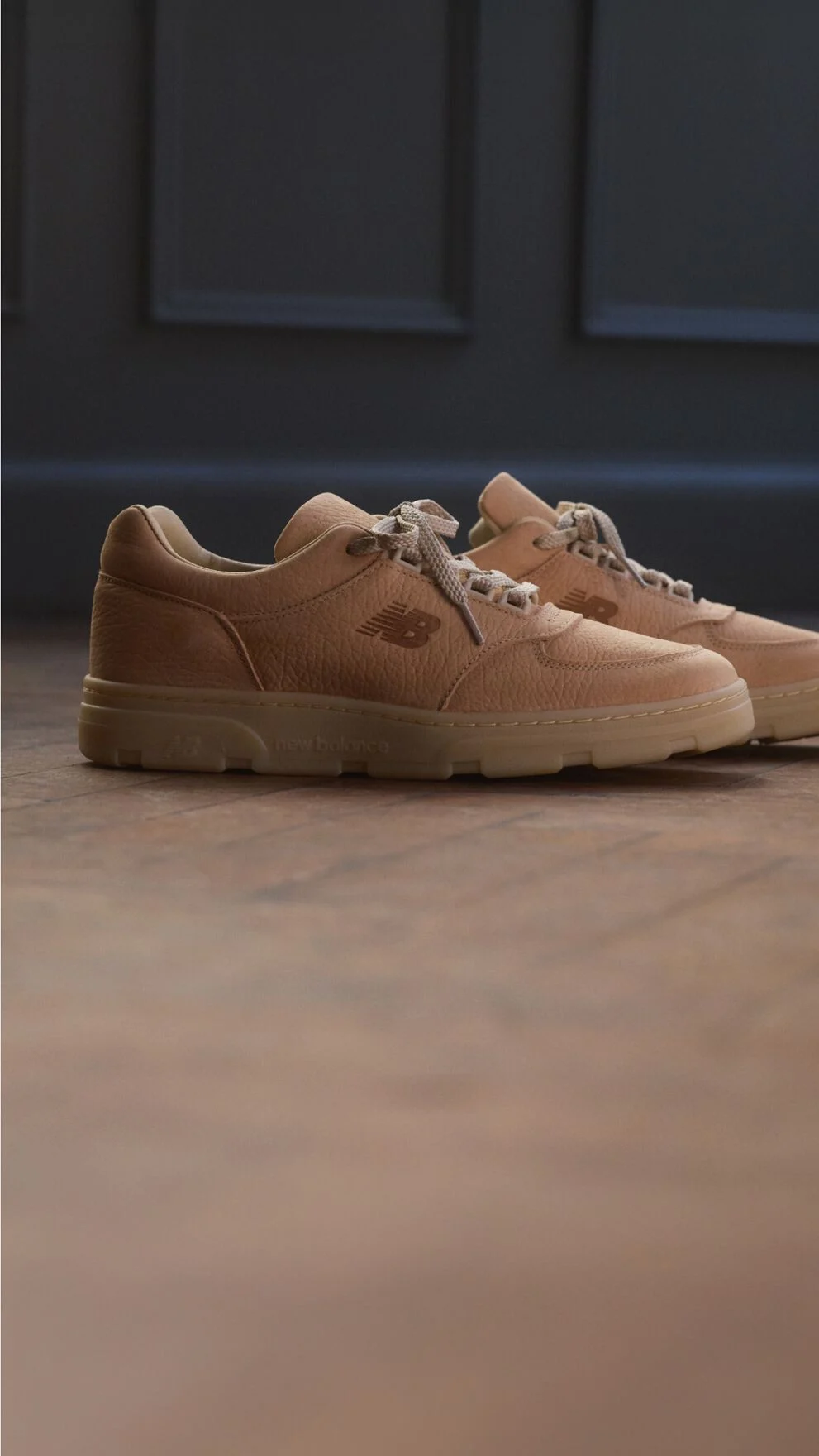 A pair of New Balance Made in UK sneakers on a wooden floor with dark paneled walls in the background.