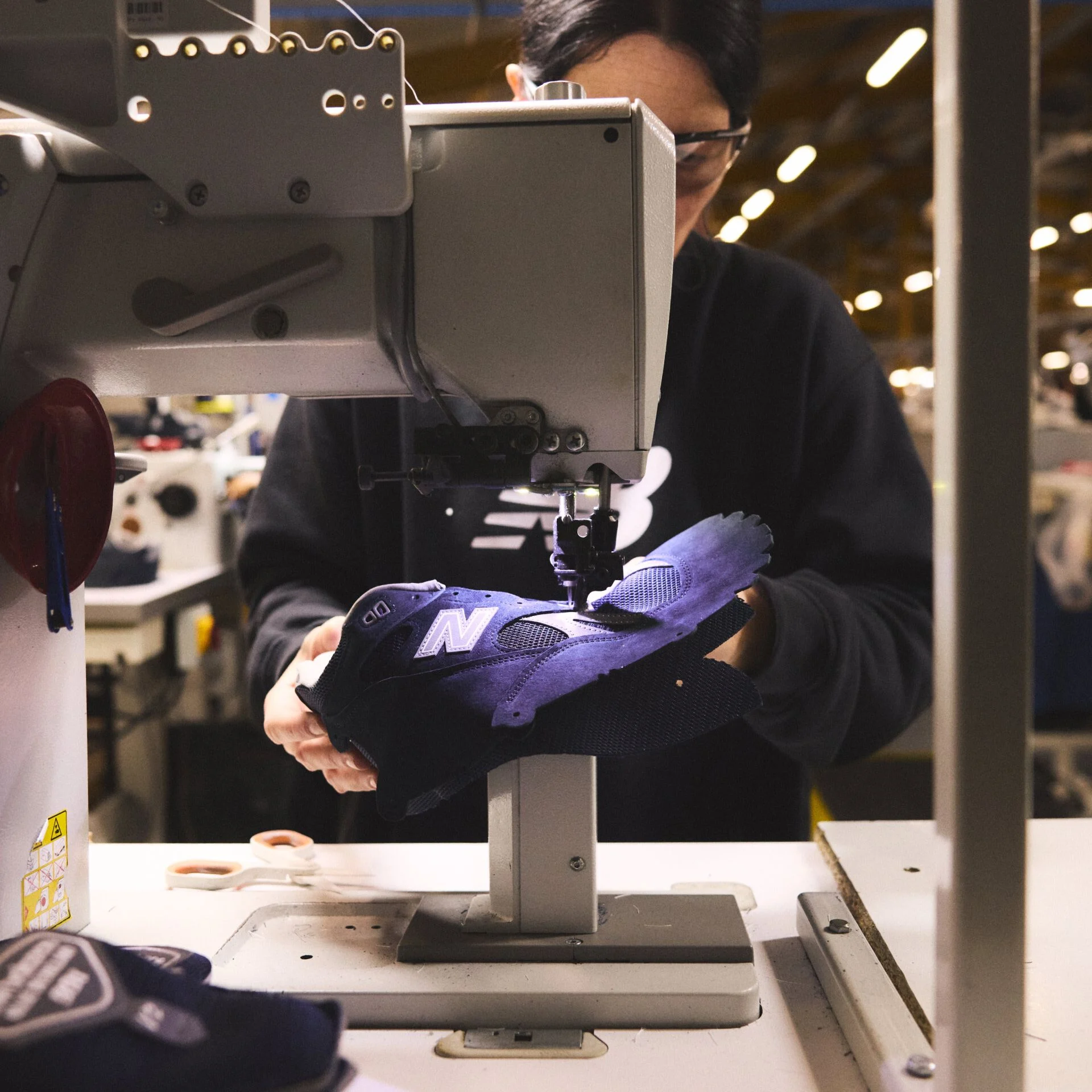 A worker in the New Balance Made in UK factory carefully sewing together a sneaker upper in an industrial environment with a sewing machine.