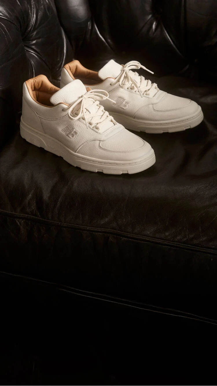 A pair of New Balance Made in UK sneakers sitting on a black leather sofa.